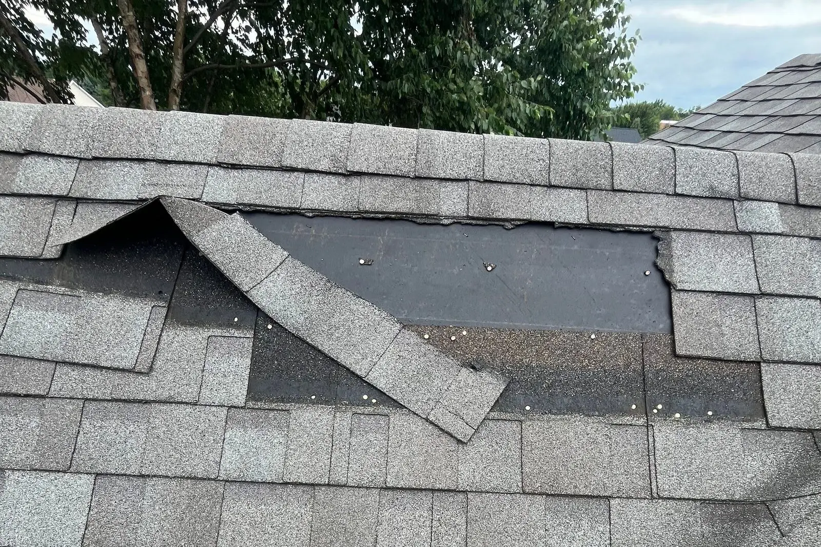 Wind-creased and torn asphalt shingles exposing the underlayment near a roof ridge