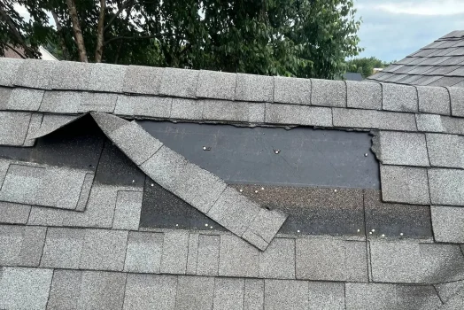 Wind-creased and torn asphalt shingles exposing the underlayment near a roof ridge